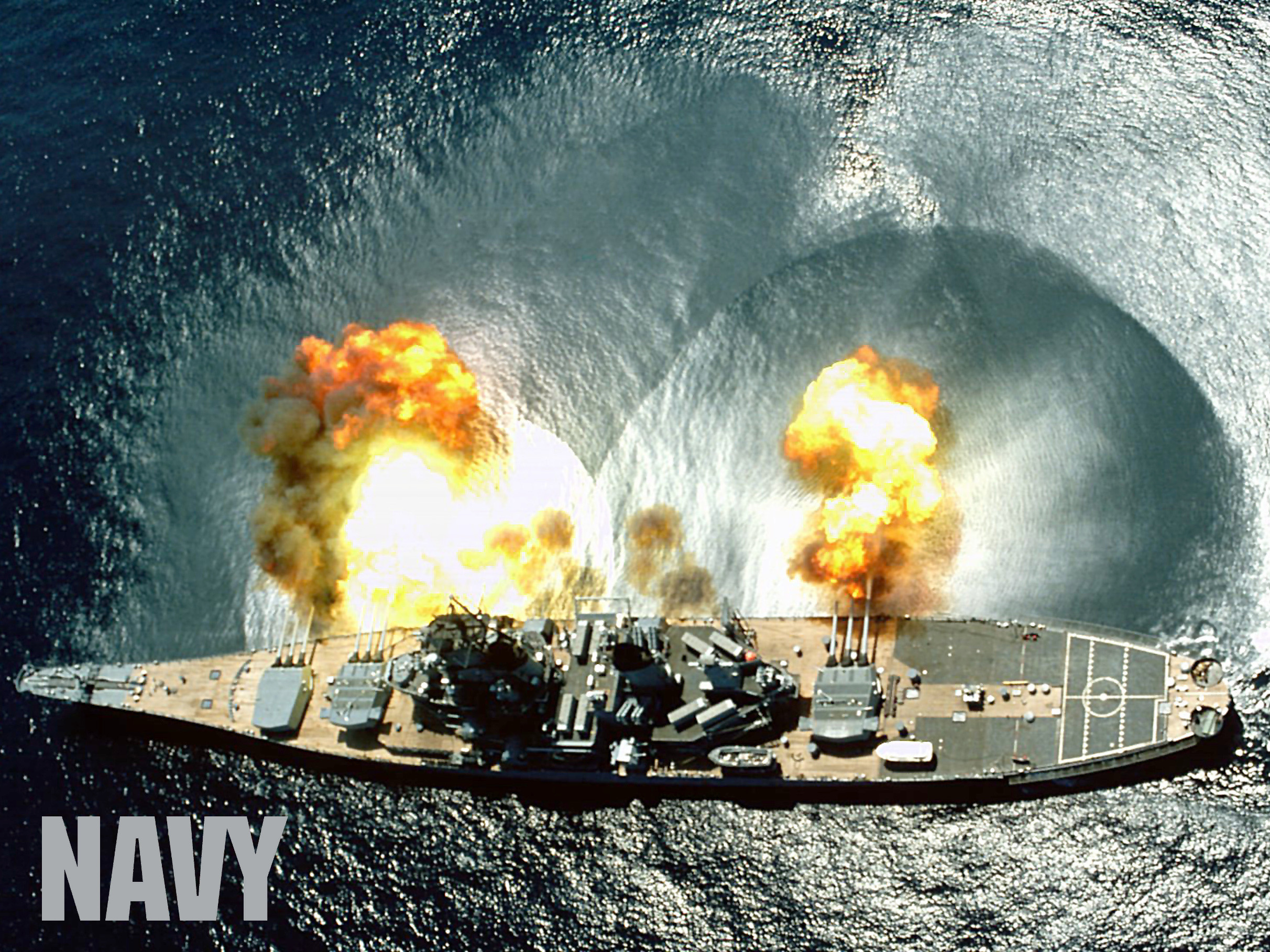 Aerial view of USS Iowa firing and the resulting shockwave on the water with the United States Navy logo type.