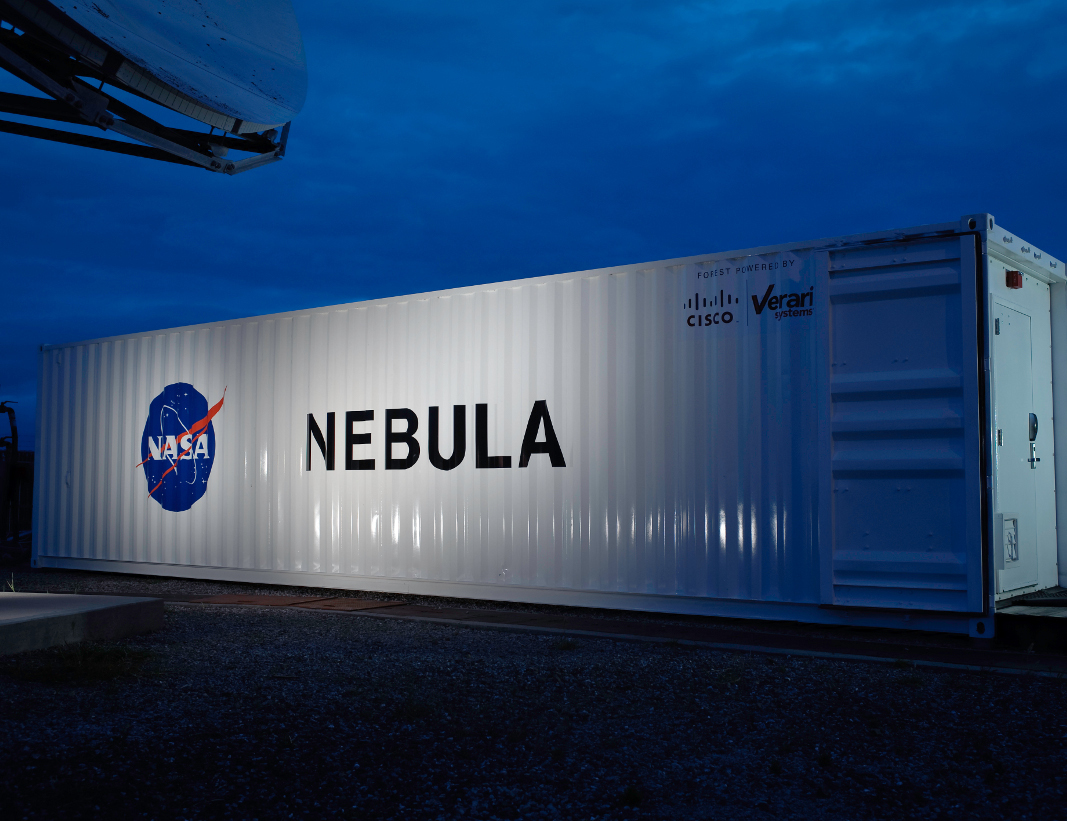Evening photograph of the NASA Nebula shipping container datacenter at dusk with splash light illuminating the Nebula logo and NASA insignia.